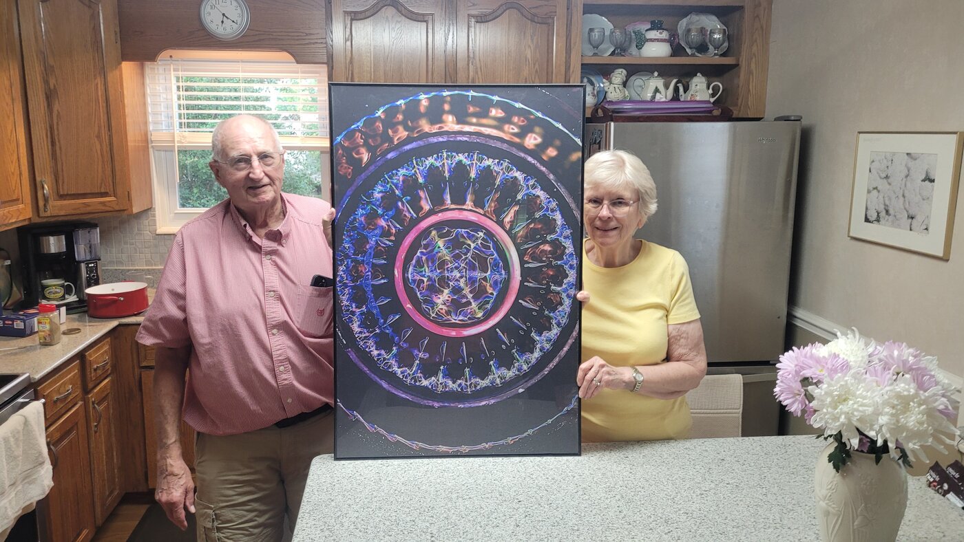 Two people holding a large framed cymatics print in a kitchen, showing its scale